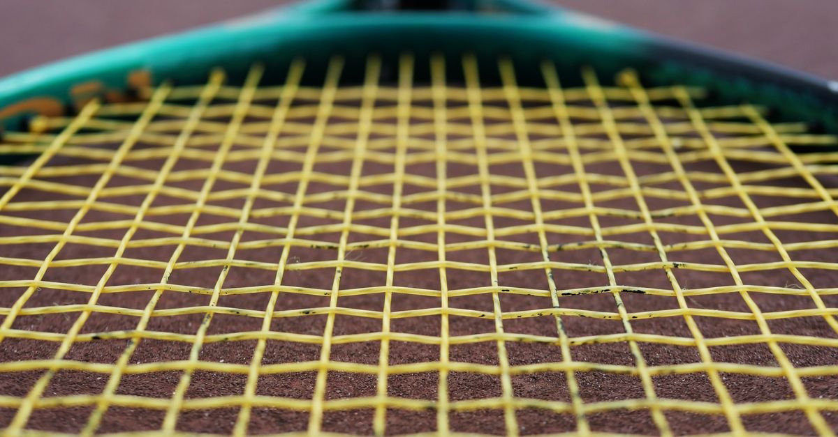 Close de raquete de tênis com encordoamento amarelo apoiada sobre quadra de saibro