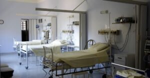 Enfermaria hospitalar com três leitos vazios, suportes de soro e equipamentos de monitoramento nas paredes