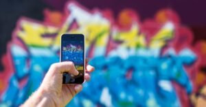 Mão segurando um telefone celular em rua com muro grafitado ao fundo