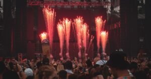 Palco de festival de música à noite com fogos de artifício e público de braços erguidos