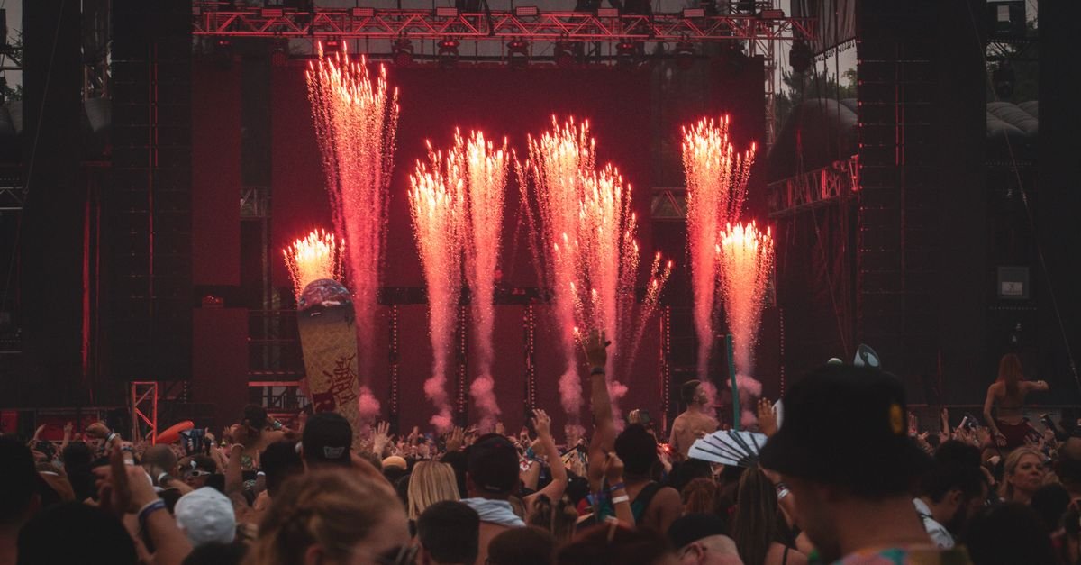 Palco de festival de música à noite com fogos de artifício e público de braços erguidos