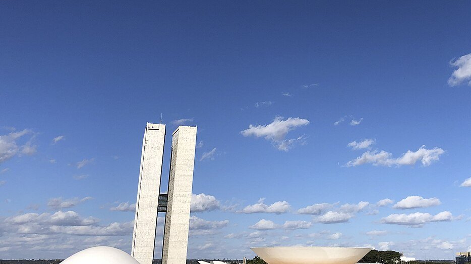 Torres do Congresso Nacional e a cúpula do plenário do Senado vistas de baixo, sob céu azul com nuvens