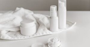 Potes e frascos brancos de dermocosméticos sobre uma superfície clara, ao lado de uma toalha
