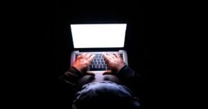 Mãos digitando em teclado de notebook em ambiente escuro, com a tela iluminada