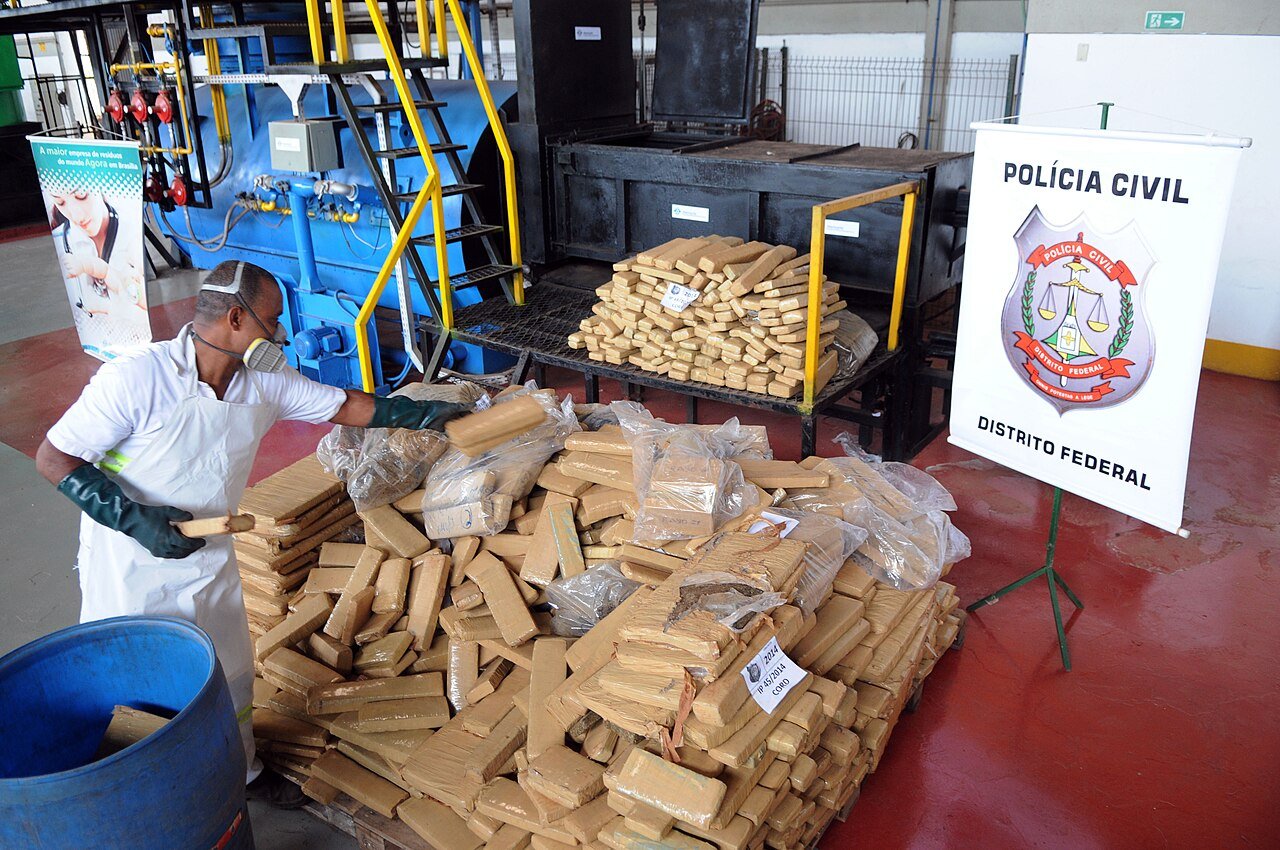Funcionário com máscara move tabletes de maconha para incineração ao lado de banner da Polícia Civil do Distrito Federal