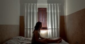 Silhueta de mulher sentada na cama diante da janela de um quarto, em contraluz