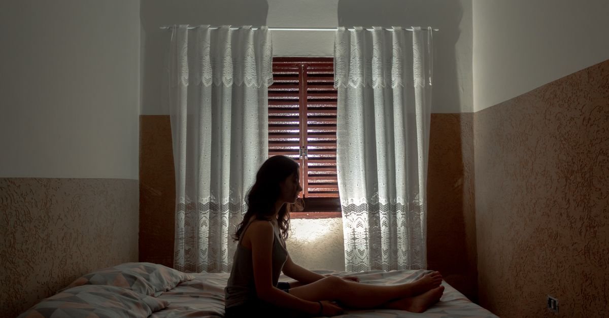 Silhueta de mulher sentada na cama diante da janela de um quarto, em contraluz