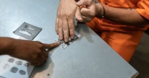 Coleta de impressões digitais de pessoa algemada sobre mesa durante procedimento de identificação criminal