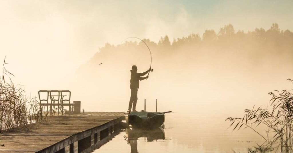 Pescador em silhueta lança a linha de uma vara de pesca em lago ao amanhecer