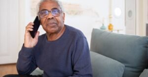 Homem idoso de oculos fala ao telefone celular sentado no sofa de casa, imagem ilustrativa
