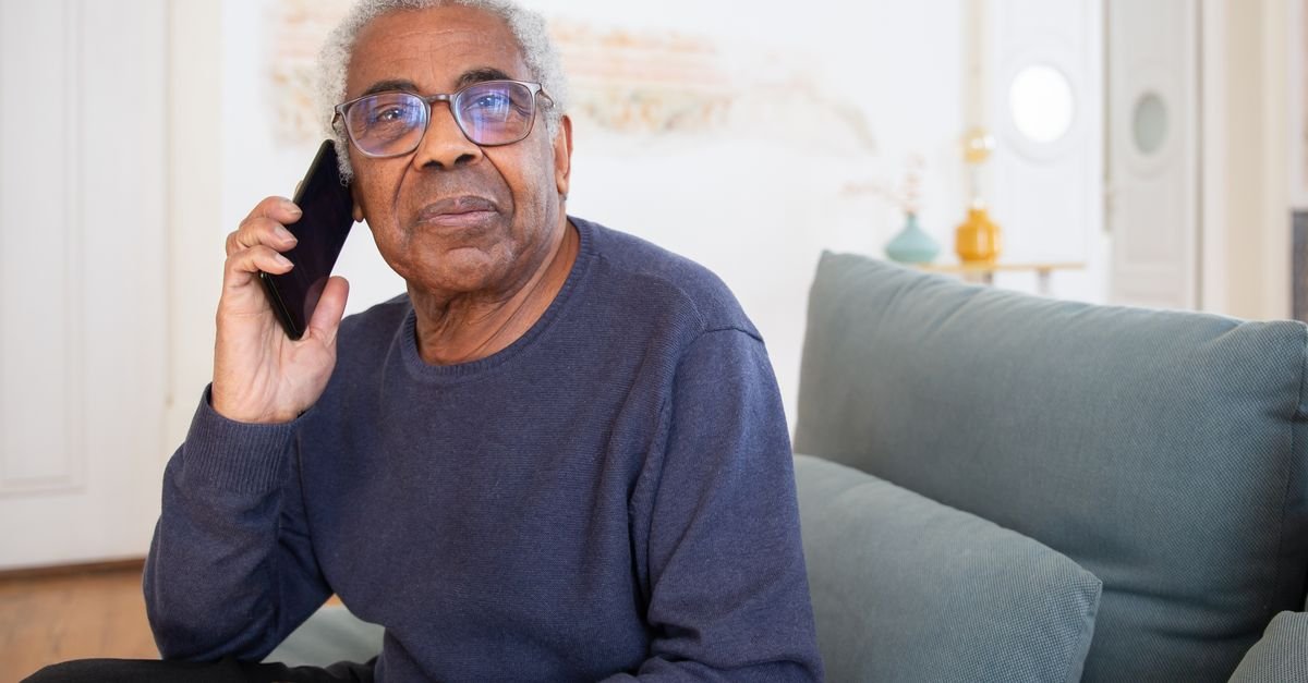 Homem idoso de oculos fala ao telefone celular sentado no sofa de casa, imagem ilustrativa