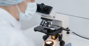 Pesquisador de jaleco, touca e máscara observa amostra em microscópio dentro de laboratório
