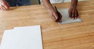 Mãos escrevendo em braile com reglete e punção sobre uma mesa de madeira