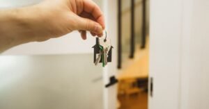 Mão segurando molho de chaves com chaveiro em formato de casa diante de porta aberta