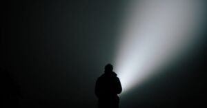 Silhueta de uma pessoa em meio à neblina, iluminada pelo facho de uma lanterna durante busca noturna