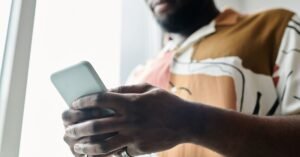 Homem segura celular junto à janela, em close das mãos sobre a tela do aparelho