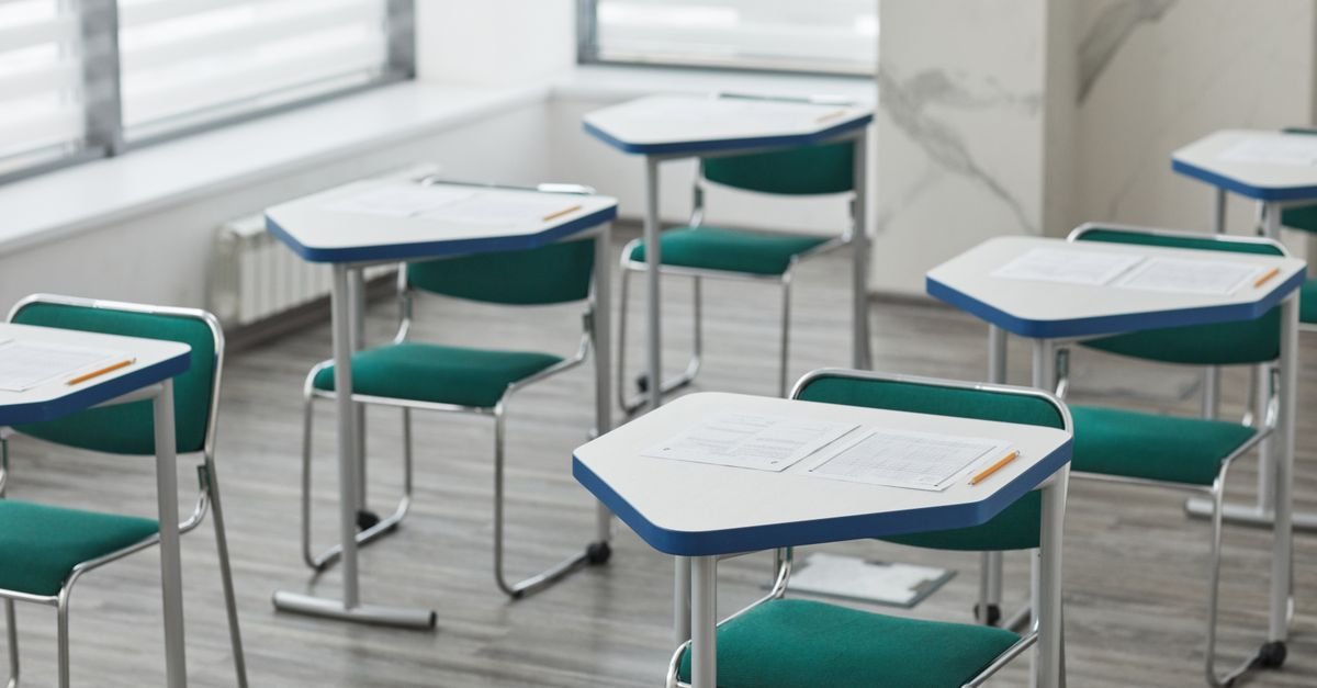 Sala de aula vazia com carteiras brancas, cadeiras verdes e folhas de exercício sobre as mesas