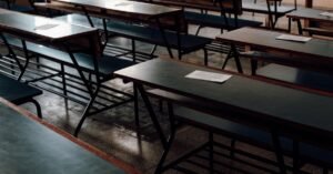Fileiras de carteiras escolares de madeira vazias em sala de aula, com folhas de papel sobre os tampos