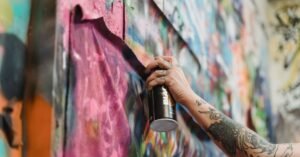 Mão segurando lata de tinta spray diante de um muro coberto de grafite colorido