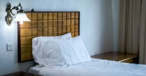 Quarto simples com cama de solteiro, cabeceira de madeira e arandela acesa na parede