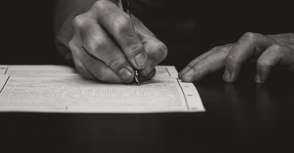 Mão de uma pessoa preenchendo um formulário impresso sobre uma mesa escura, com caneta
