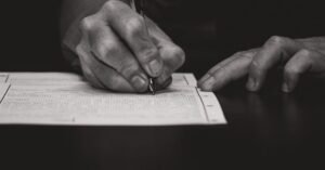 Mão de uma pessoa preenchendo um formulário impresso sobre uma mesa escura, com caneta