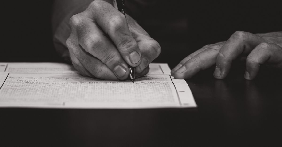 Mão de uma pessoa preenchendo um formulário impresso sobre uma mesa escura, com caneta