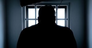 Silhueta de homem de costas diante de janela com grades em ambiente escuro