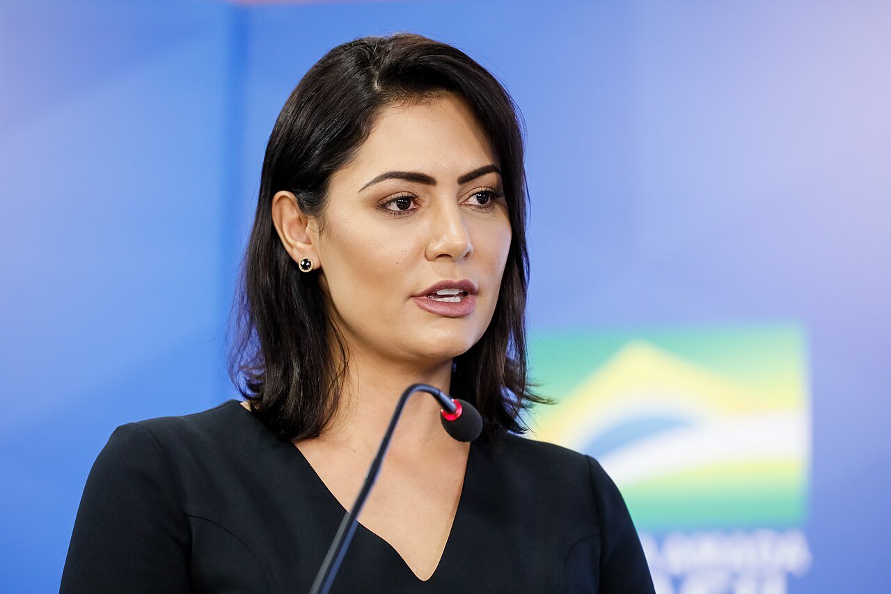 Michelle Bolsonaro fala ao microfone diante de um painel azul com a bandeira do Brasil ao fundo