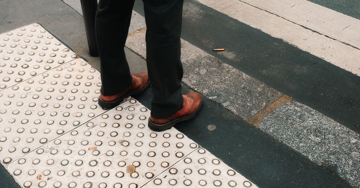 Pedestre parado sobre piso tátil de calçada ao lado de faixa de travessia em via urbana
