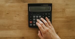 Mão de uma pessoa apertando as teclas de uma calculadora preta sobre mesa de madeira
