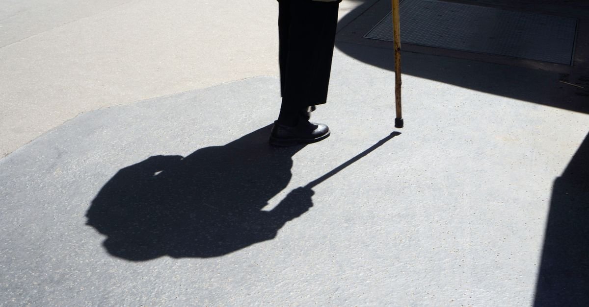 Sombra projetada no chão de pessoa idosa caminhando apoiada em bengala, em calçada iluminada pelo sol