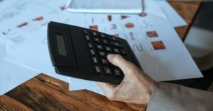 Mão de uma pessoa operando uma calculadora sobre mesa de madeira com planilhas e gráficos impressos