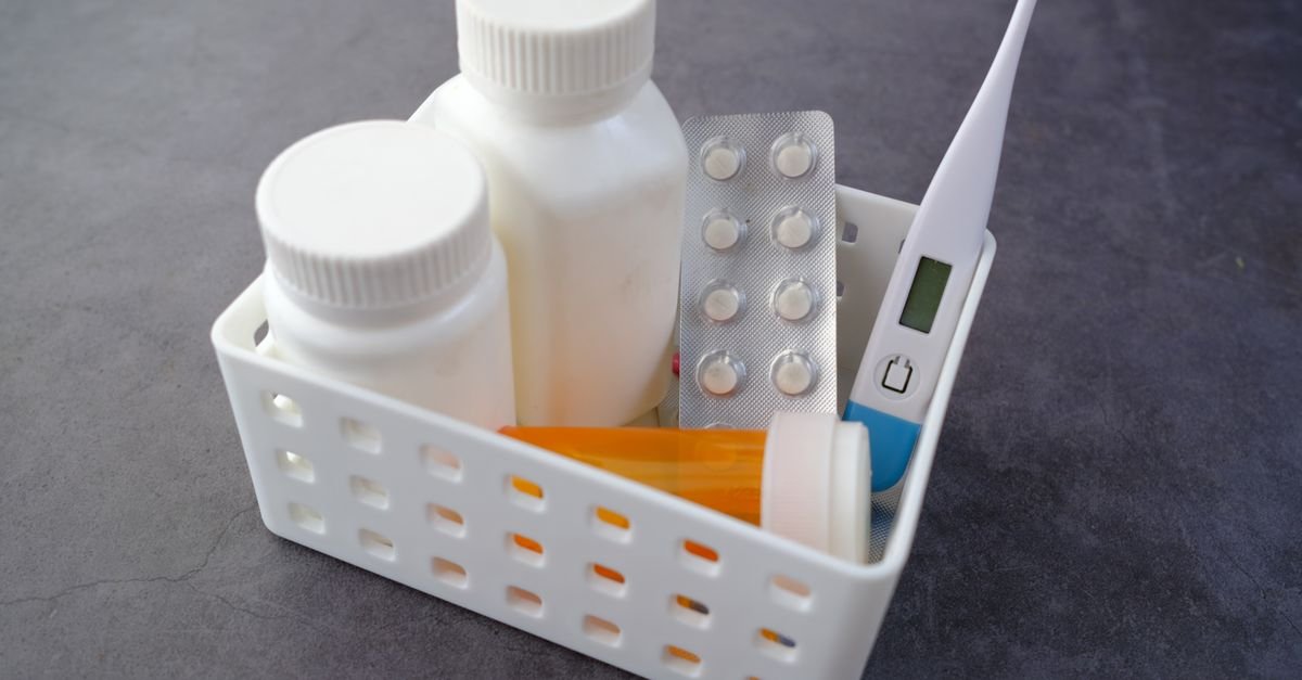 Cesta branca com frascos de medicamento, cartela de comprimidos e termômetro sobre superfície escura