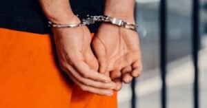 Maos de um homem algemadas atras do corpo, em closeup, com uniforme laranja de detento ao fundo