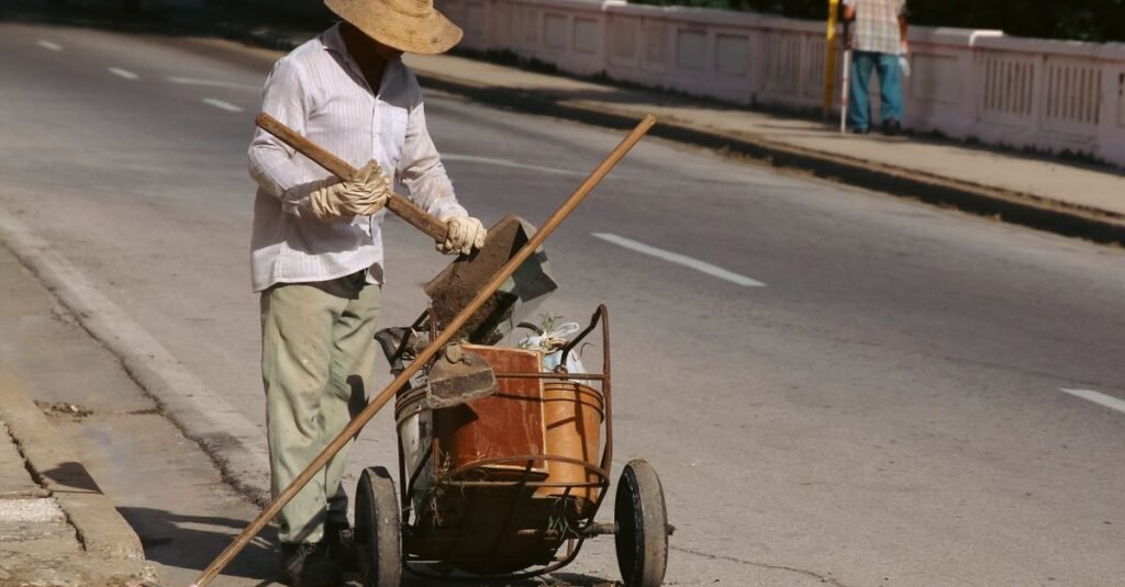 Trabalhador de chapéu recolhe resíduos da via sob sol forte, ao lado de carrinho de coleta