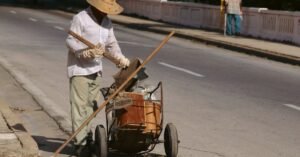 Trabalhador de chapéu recolhe resíduos da via sob sol forte, ao lado de carrinho de coleta