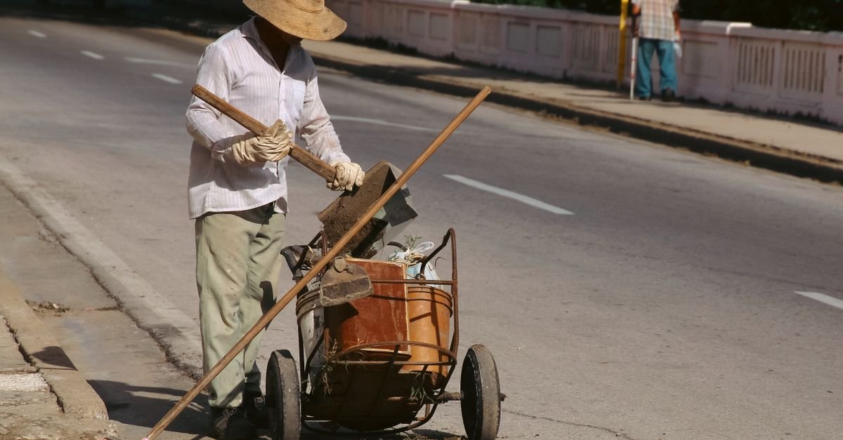 Trabalhador de chapéu recolhe resíduos da via sob sol forte, ao lado de carrinho de coleta
