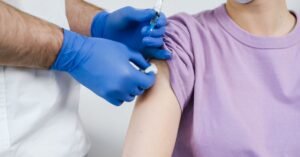 Profissional de saude com luvas azuis aplica vacina no braco de um paciente