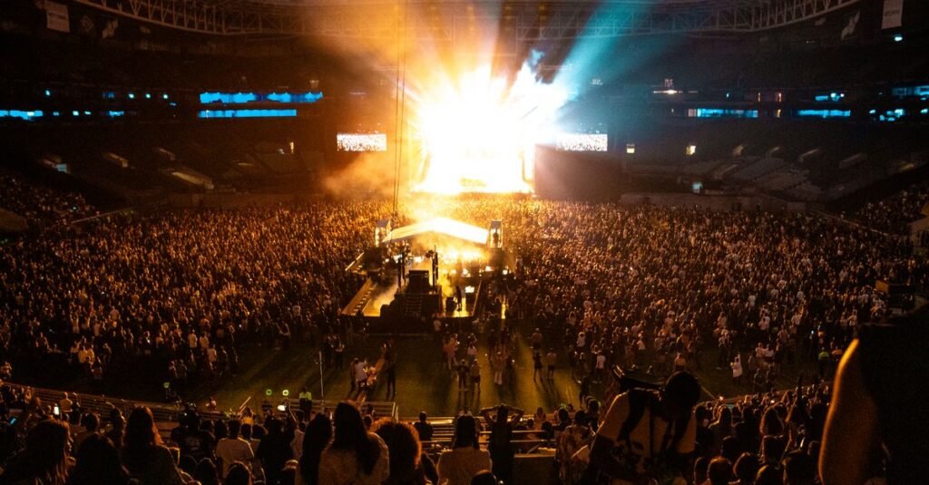 Estádio lotado durante show noturno, com feixes de luz saindo do palco sobre o público no gramado e nas arquibancadas
