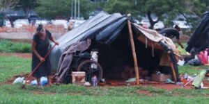 Barraca de lona armada em área verde, com pertences ao redor, e mulher varrendo o terreno ao lado