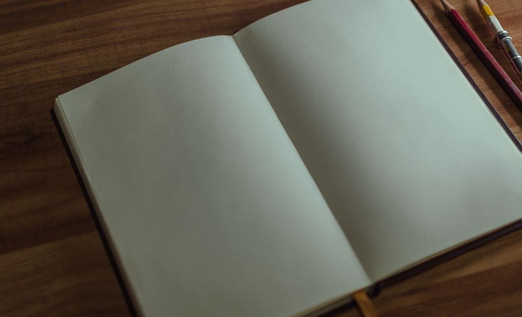 Caderno aberto em branco sobre mesa de madeira, ao lado de dois lápis