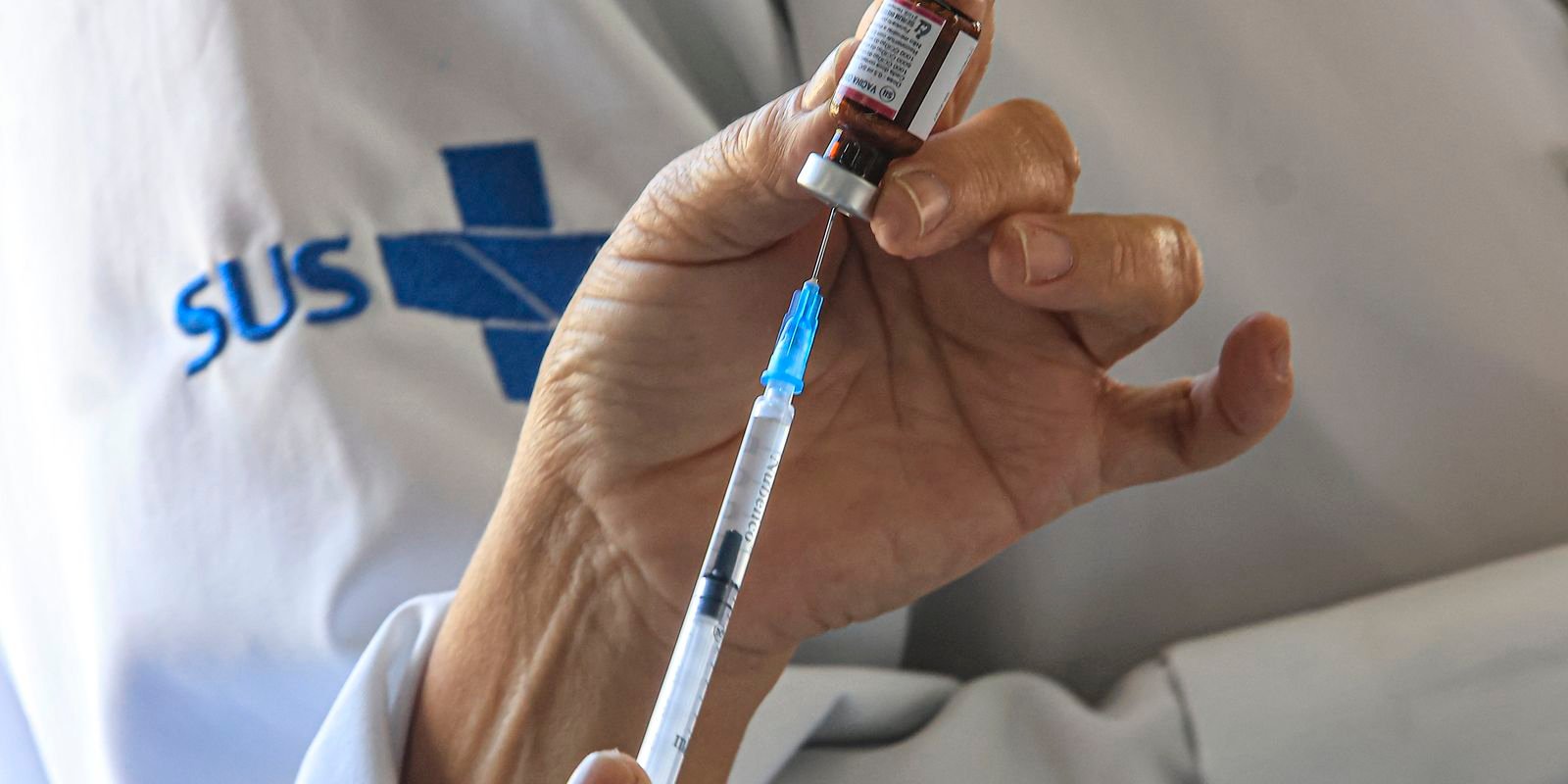 Profissional de saude com jaleco do SUS prepara uma seringa a partir de um frasco de vacina