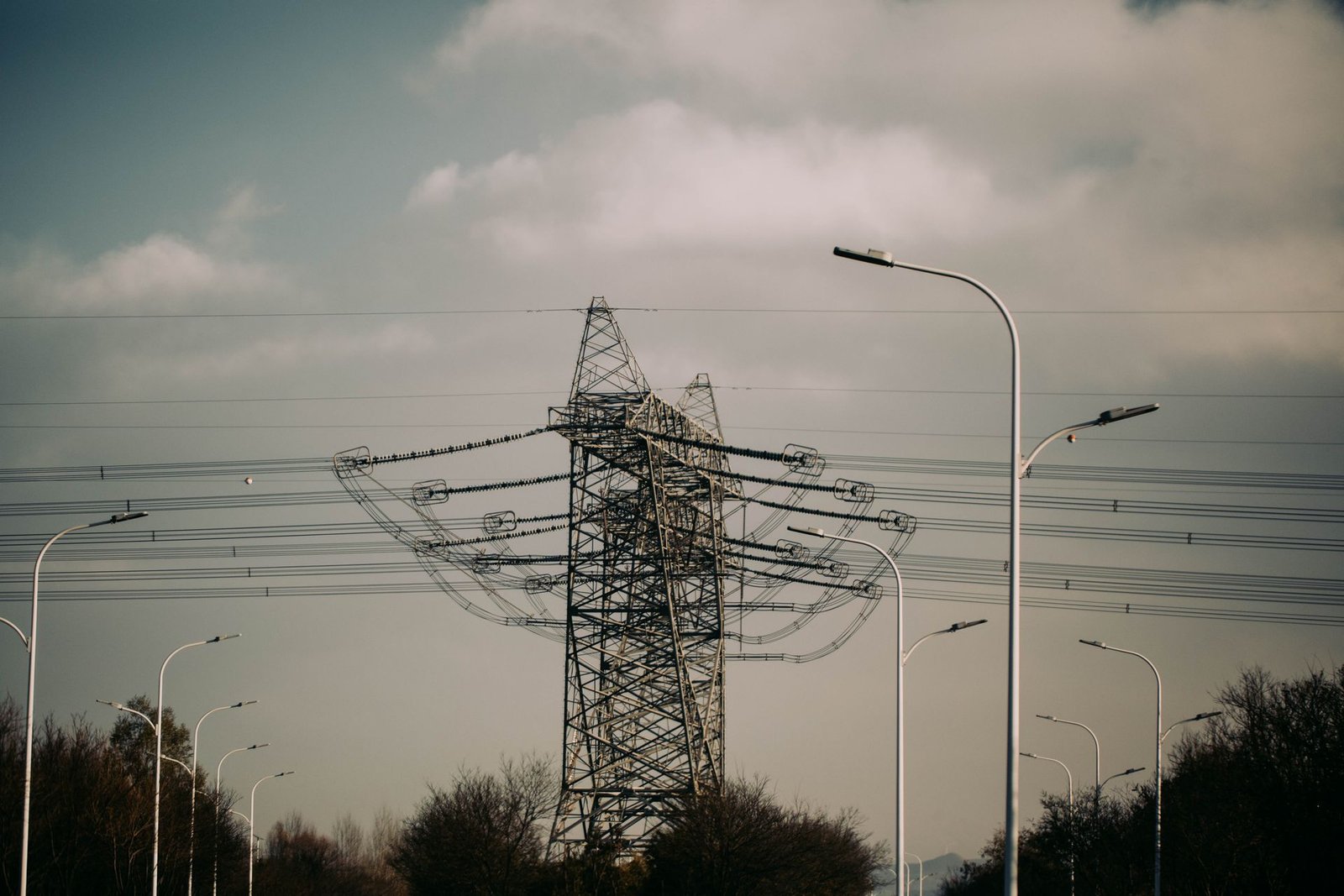 Torre de transmissão de alta tensão com linhas de energia e postes de iluminação ao fundo, sob céu nublado