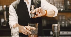 Barman prepara bebida atrás do balcão de um bar, com copos e dosador sobre a bancada