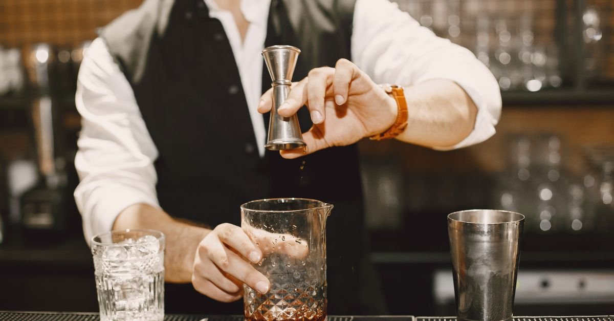 Barman prepara bebida atrás do balcão de um bar, com copos e dosador sobre a bancada