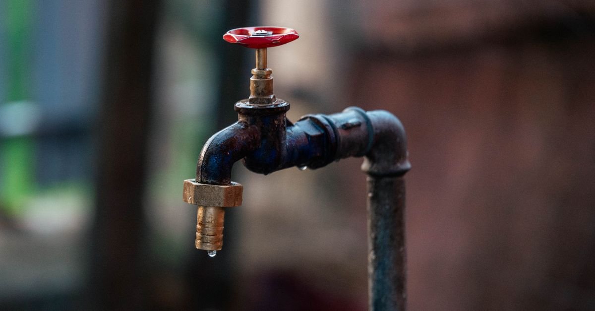 Torneira antiga de metal com registro vermelho e uma gota d'água caindo do bico