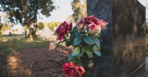 Buquê de rosas em vaso metálico apoiado em lápide de granito escuro, com cemitério desfocado ao fundo