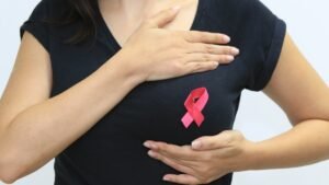 Mulher com laço rosa na blusa apalpa a própria mama em gesto de autoexame