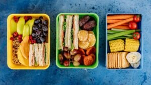 Três lancheiras escolares abertas com frutas, sanduíches e legumes sobre mesa azul